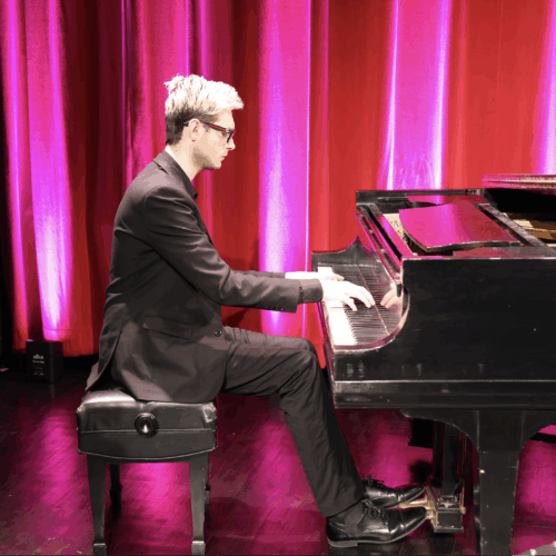 Joseph Toto playing a black grand piano at the PAC with red curtain behind him.